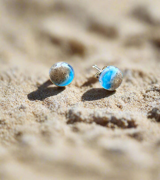 This is a picture of two small round earrings laying on beach sand.