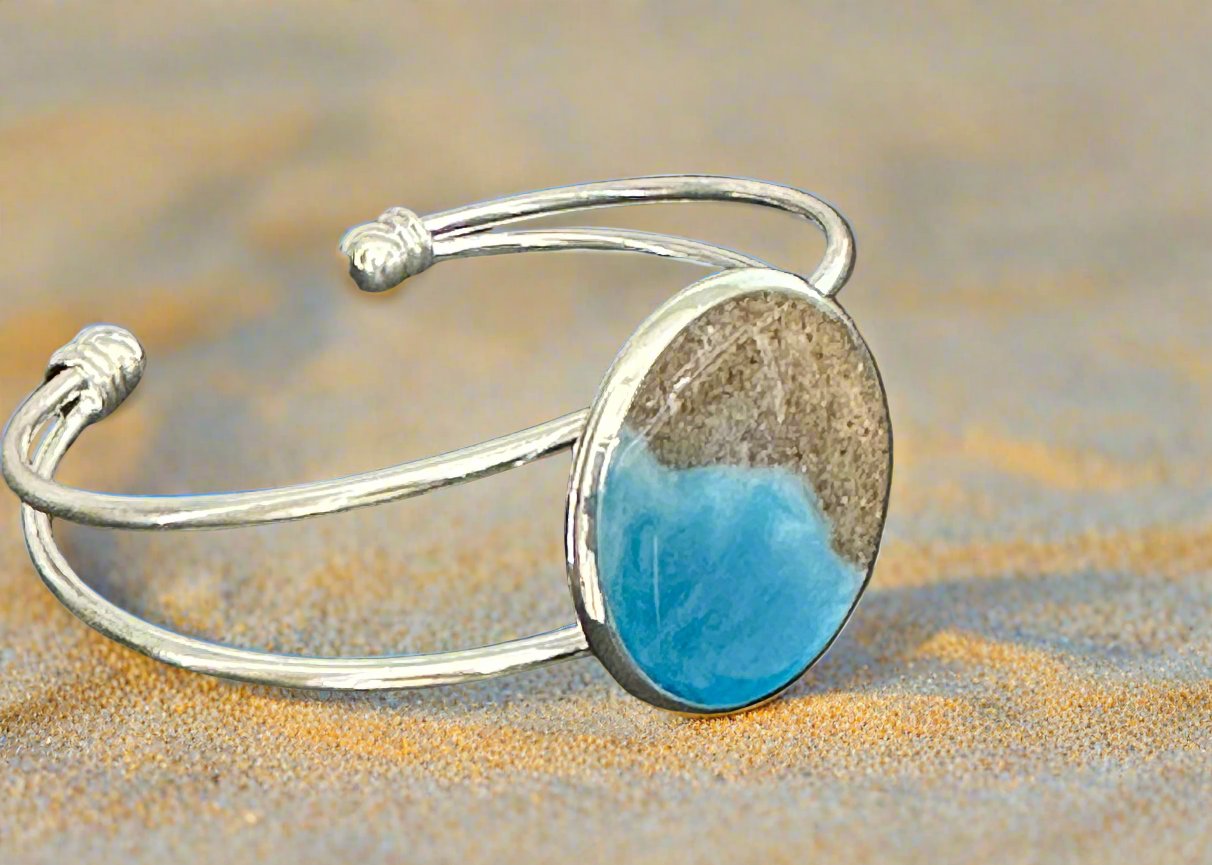 This is a picture of a silver bangle bracelet with round bezel in the center. The bezel has sand and resin in the shape of the coast.