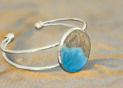 This is a picture of a silver bangle bracelet with round bezel in the center. The bezel has sand and resin in the shape of the coast.