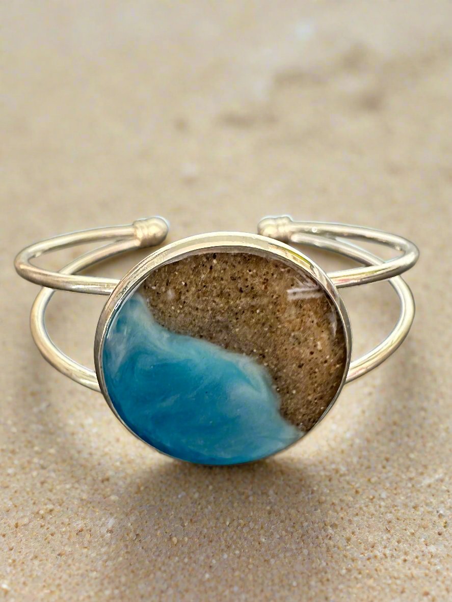 A silver bangle bracelet with a bezel in the center. Round bezel is filled with sand and resin in the shape of the coastline.